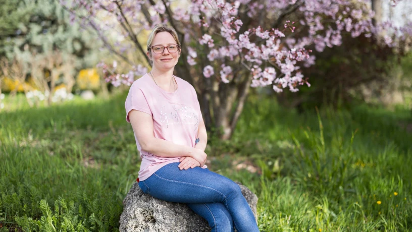 Bild einer jungen Frau auf einem Stein sitzend vor einem Garten.